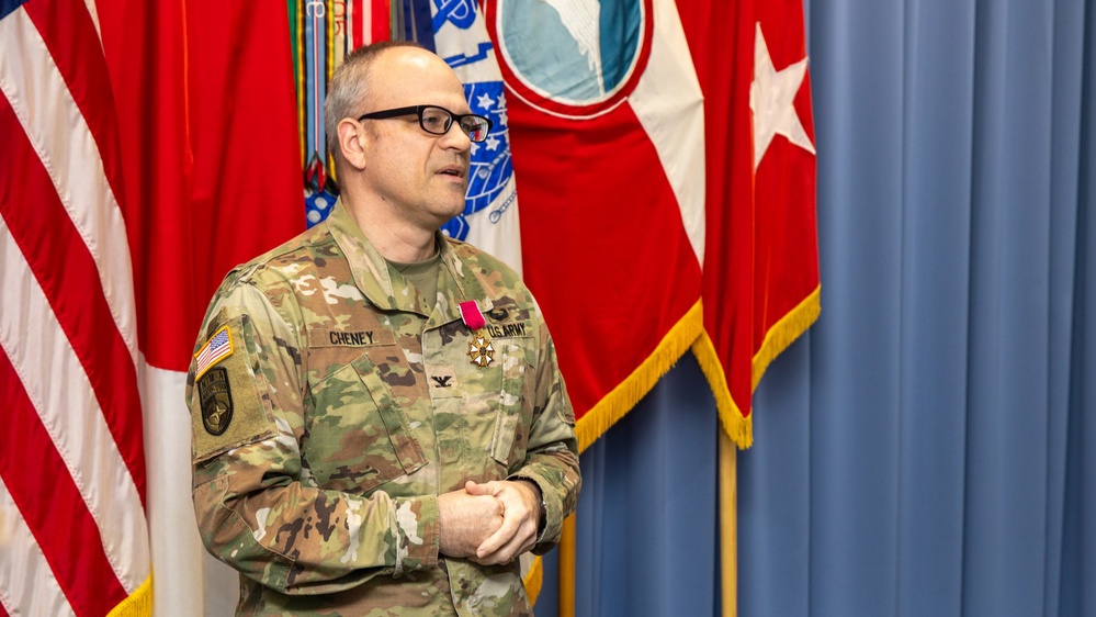 A U.S. Army Colonel named Cheney stands before a display of military flags, wearing an OCP (Operational Camouflage Pattern) uniform with a medal pinned to his chest. He appears to be speaking or addressing an audience during what appears to be a formal military ceremony. Multiple flags including the U.S. flag and what appears to be a general officer flag are visible in the background.