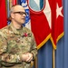 A U.S. Army Colonel named Cheney stands before a display of military flags, wearing an OCP (Operational Camouflage Pattern) uniform with a medal pinned to his chest. He appears to be speaking or addressing an audience during what appears to be a formal military ceremony. Multiple flags including the U.S. flag and what appears to be a general officer flag are visible in the background.