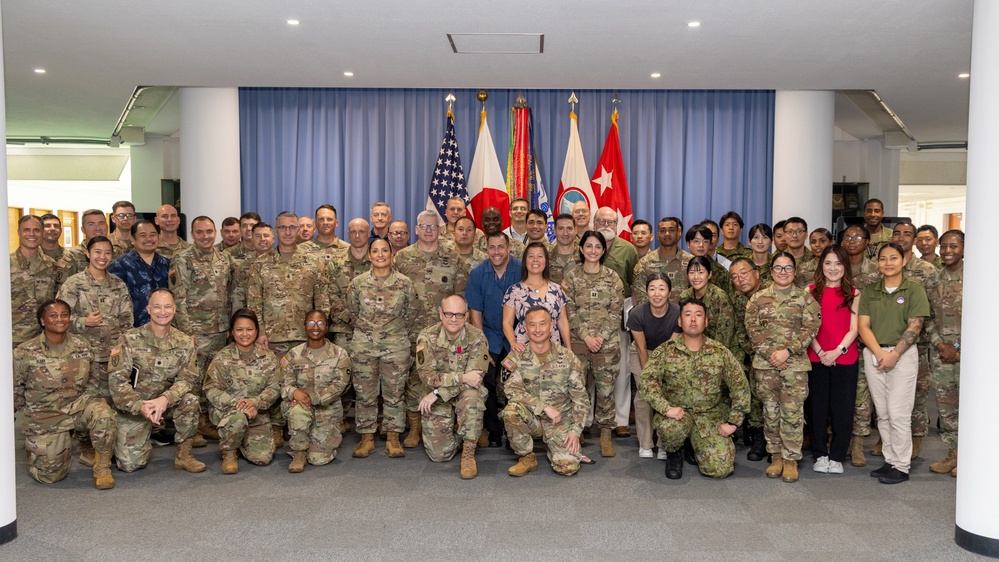 A large group photograph of approximately 60-70 military personnel and civilians posing together in a formal indoor setting. The group includes U.S. Army soldiers in OCP (Operational Camouflage Pattern) uniforms, Japanese Self-Defense Force members, and civilian personnel. Multiple national and military flags are displayed in the background, suggesting a joint military event or ceremony.
