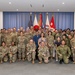 A large group photograph of approximately 60-70 military personnel and civilians posing together in a formal indoor setting. The group includes U.S. Army soldiers in OCP (Operational Camouflage Pattern) uniforms, Japanese Self-Defense Force members, and civilian personnel. Multiple national and military flags are displayed in the background, suggesting a joint military event or ceremony.