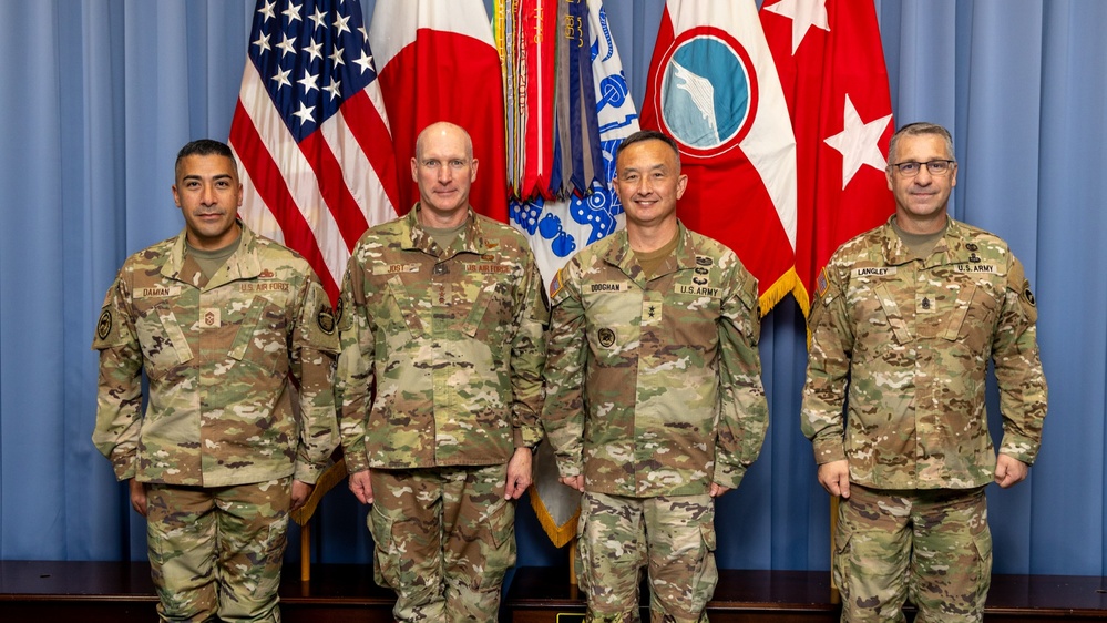 Four military personnel in OCP (Operational Camouflage Pattern) uniforms pose for a formal group photograph in front of multiple flags. The individuals represent both U.S. Air Force and U.S. Army branches, identifiable by name tapes reading DAMIAN, JOST, DOOCHAN, and LANGLEY. The setting appears to be an official military headquarters or command facility.