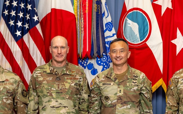 Four military personnel in OCP (Operational Camouflage Pattern) uniforms pose for a formal group photograph in front of multiple flags. The individuals represent both U.S. Air Force and U.S. Army branches, identifiable by name tapes reading DAMIAN, JOST, DOOCHAN, and LANGLEY. The setting appears to be an official military headquarters or command facility.