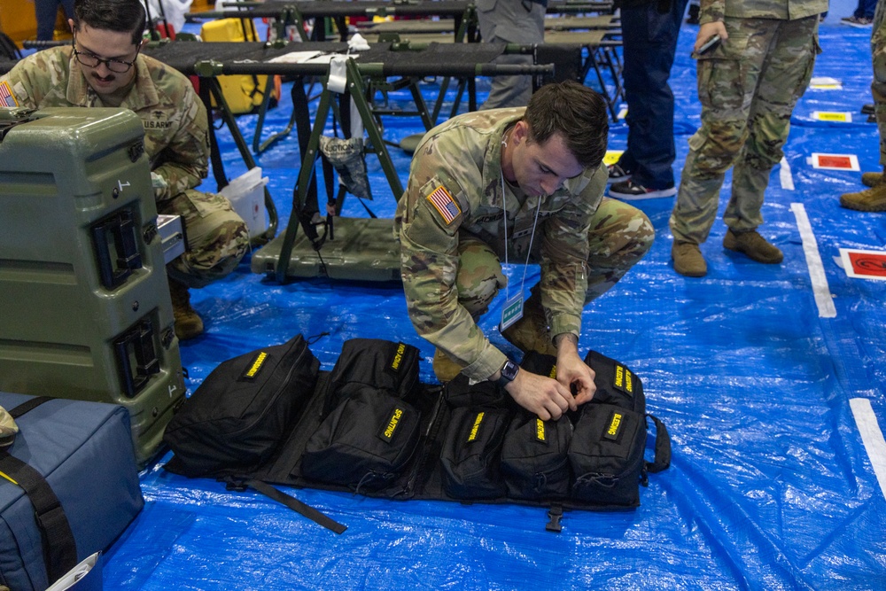 U.S. Army soldiers are setting up or organizing equipment in what appears to be a large indoor facility with blue tarp flooring. The foreground soldier is crouching over multiple black bags with yellow text labeling, while a second soldier works with military cases in the background. The setting suggests a field hospital setup, emergency deployment, or military exercise staging area.