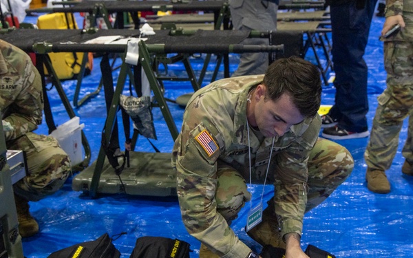 U.S. Army soldiers are setting up or organizing equipment in what appears to be a large indoor facility with blue tarp flooring. The foreground soldier is crouching over multiple black bags with yellow text labeling, while a second soldier works with military cases in the background. The setting suggests a field hospital setup, emergency deployment, or military exercise staging area.