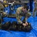 U.S. Army soldiers are setting up or organizing equipment in what appears to be a large indoor facility with blue tarp flooring. The foreground soldier is crouching over multiple black bags with yellow text labeling, while a second soldier works with military cases in the background. The setting suggests a field hospital setup, emergency deployment, or military exercise staging area.