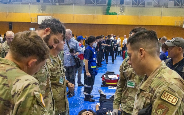 A large-scale joint military and civilian medical exercise is taking place inside a gymnasium or sports hall. U.S. military personnel in multicam/OCP uniforms, including at least one with a 'MED' patch indicating medical specialty, are attending to simulated casualties on stretchers. The exercise appears to involve multinational participants including Japanese civilian emergency responders and military personnel.