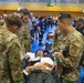 A large-scale joint military and civilian medical exercise is taking place inside a gymnasium or sports hall. U.S. military personnel in multicam/OCP uniforms, including at least one with a 'MED' patch indicating medical specialty, are attending to simulated casualties on stretchers. The exercise appears to involve multinational participants including Japanese civilian emergency responders and military personnel.