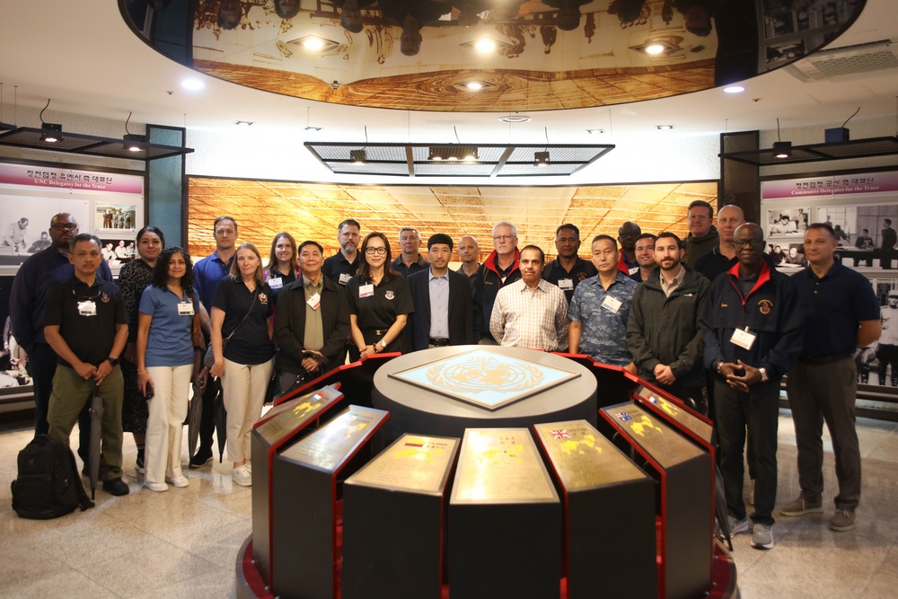 A group of approximately 20-25 individuals, appearing to be military personnel and civilians, pose for a group photo inside what appears to be a Korean War memorial museum or exhibition hall. The central display features a circular podium with the United Nations emblem and plaques representing contributing nations. Korean text and exhibition panels are visible in the background.