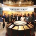 A group of approximately 20-25 individuals, appearing to be military personnel and civilians, pose for a group photo inside what appears to be a Korean War memorial museum or exhibition hall. The central display features a circular podium with the United Nations emblem and plaques representing contributing nations. Korean text and exhibition panels are visible in the background.