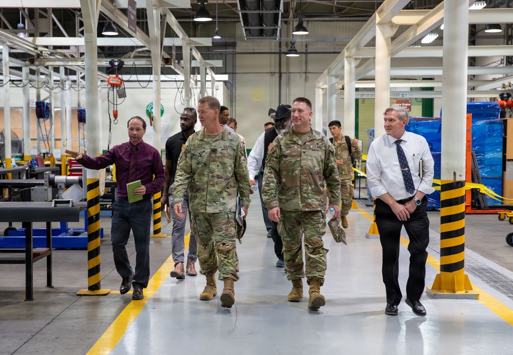A group of individuals, including two U.S. Army soldiers in OCP (Operational Camouflage Pattern) uniforms, are walking through what appears to be a military maintenance or industrial facility. They are accompanied by civilians, one of whom appears to be gesturing and explaining something, suggesting this is an official tour or inspection. The facility features industrial equipment, overhead hoists, and safety-marked bollards.