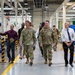 A group of individuals, including two U.S. Army soldiers in OCP (Operational Camouflage Pattern) uniforms, are walking through what appears to be a military maintenance or industrial facility. They are accompanied by civilians, one of whom appears to be gesturing and explaining something, suggesting this is an official tour or inspection. The facility features industrial equipment, overhead hoists, and safety-marked bollards.