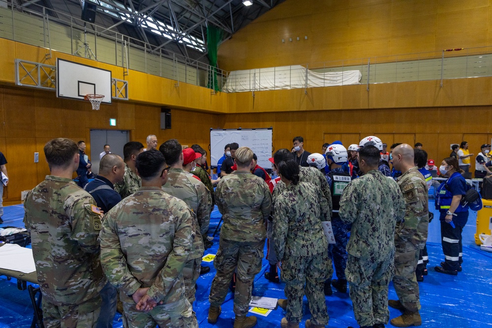 A joint military and emergency response briefing is taking place inside a gymnasium, involving U.S. Army soldiers in OCP camouflage, U.S. Navy personnel in NWU camouflage, and Japanese emergency responders wearing white helmets and blue uniforms. The group is gathered around a whiteboard in what appears to be a coordinated disaster response or humanitarian assistance exercise. The setting suggests a multinational training event or real-world emergency operation conducted in Japan.