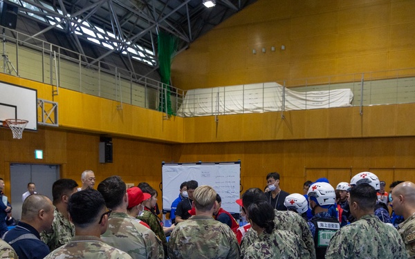 A joint military and emergency response briefing is taking place inside a gymnasium, involving U.S. Army soldiers in OCP camouflage, U.S. Navy personnel in NWU camouflage, and Japanese emergency responders wearing white helmets and blue uniforms. The group is gathered around a whiteboard in what appears to be a coordinated disaster response or humanitarian assistance exercise. The setting suggests a multinational training event or real-world emergency operation conducted in Japan.
