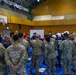 A joint military and emergency response briefing is taking place inside a gymnasium, involving U.S. Army soldiers in OCP camouflage, U.S. Navy personnel in NWU camouflage, and Japanese emergency responders wearing white helmets and blue uniforms. The group is gathered around a whiteboard in what appears to be a coordinated disaster response or humanitarian assistance exercise. The setting suggests a multinational training event or real-world emergency operation conducted in Japan.