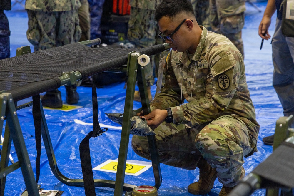 A U.S. Army soldier wearing OCP (Operational Camouflage Pattern) uniform with a Medical Corps insignia is crouching down and assembling or adjusting a military field cot/litter on a blue tarp floor. The soldier's name tape appears to read 'PITAIT' and the branch tape reads 'ARMY'. The setting appears to be an indoor military medical training or setup exercise, with Navy personnel visible in the background.