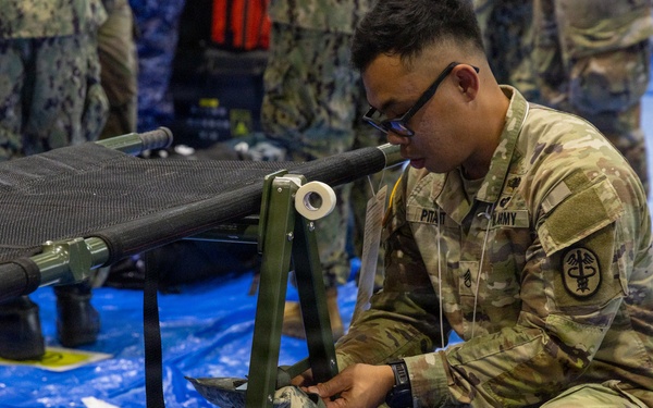 A U.S. Army soldier wearing OCP (Operational Camouflage Pattern) uniform with a Medical Corps insignia is crouching down and assembling or adjusting a military field cot/litter on a blue tarp floor. The soldier's name tape appears to read 'PITAIT' and the branch tape reads 'ARMY'. The setting appears to be an indoor military medical training or setup exercise, with Navy personnel visible in the background.