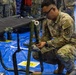 A U.S. Army soldier wearing OCP (Operational Camouflage Pattern) uniform with a Medical Corps insignia is crouching down and assembling or adjusting a military field cot/litter on a blue tarp floor. The soldier's name tape appears to read 'PITAIT' and the branch tape reads 'ARMY'. The setting appears to be an indoor military medical training or setup exercise, with Navy personnel visible in the background.