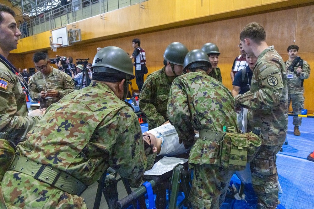 Military personnel from what appear to be two different armed forces — U.S. Army soldiers in MultiCam/OCP uniforms and soldiers in Japanese Ground Self-Defense Force uniforms with distinctive helmets — are conducting a joint medical training exercise inside a gymnasium. They are gathered around a stretcher with a simulated casualty, reviewing documents and practicing medical procedures. Media personnel with cameras are visible in the background, suggesting this is a documented joint exercise or drill.