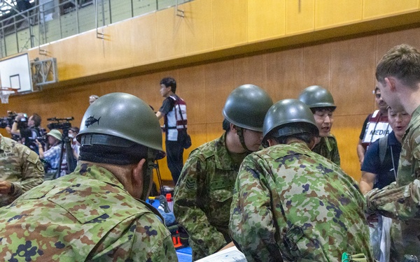 Military personnel from what appear to be two different armed forces — U.S. Army soldiers in MultiCam/OCP uniforms and soldiers in Japanese Ground Self-Defense Force uniforms with distinctive helmets — are conducting a joint medical training exercise inside a gymnasium. They are gathered around a stretcher with a simulated casualty, reviewing documents and practicing medical procedures. Media personnel with cameras are visible in the background, suggesting this is a documented joint exercise or drill.