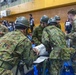 Military personnel from what appear to be two different armed forces — U.S. Army soldiers in MultiCam/OCP uniforms and soldiers in Japanese Ground Self-Defense Force uniforms with distinctive helmets — are conducting a joint medical training exercise inside a gymnasium. They are gathered around a stretcher with a simulated casualty, reviewing documents and practicing medical procedures. Media personnel with cameras are visible in the background, suggesting this is a documented joint exercise or drill.