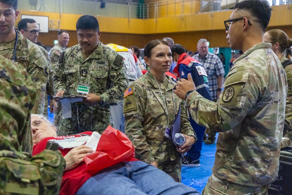 The image depicts a military medical exercise or disaster response drill taking place inside a gymnasium. Personnel from the U.S. Army and U.S. Navy are gathered around a simulated casualty lying on a stretcher, appearing to coordinate triage and medical response activities. Civilian participants and observers are also present in the background, suggesting a joint or multi-agency training event.