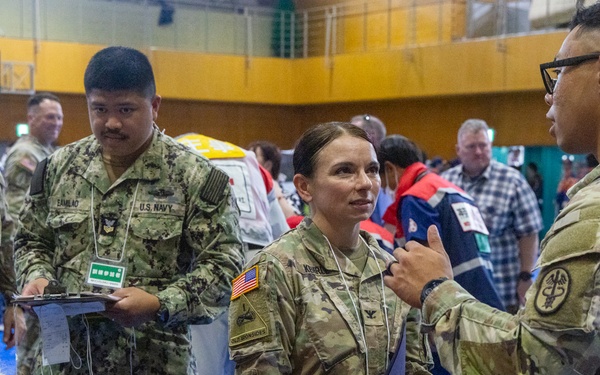 The image depicts a military medical exercise or disaster response drill taking place inside a gymnasium. Personnel from the U.S. Army and U.S. Navy are gathered around a simulated casualty lying on a stretcher, appearing to coordinate triage and medical response activities. Civilian participants and observers are also present in the background, suggesting a joint or multi-agency training event.