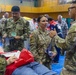 The image depicts a military medical exercise or disaster response drill taking place inside a gymnasium. Personnel from the U.S. Army and U.S. Navy are gathered around a simulated casualty lying on a stretcher, appearing to coordinate triage and medical response activities. Civilian participants and observers are also present in the background, suggesting a joint or multi-agency training event.