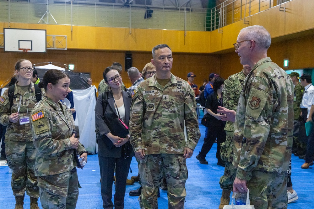 A group of U.S. military personnel in OCP (Operational Camouflage Pattern) uniforms are engaged in conversation inside what appears to be a gymnasium or community center being used as an operational or humanitarian assistance facility. At least three branches appear to be represented, including U.S. Army and U.S. Air Force personnel. A civilian woman in business attire is also present among the group, suggesting a joint civil-military coordination meeting.