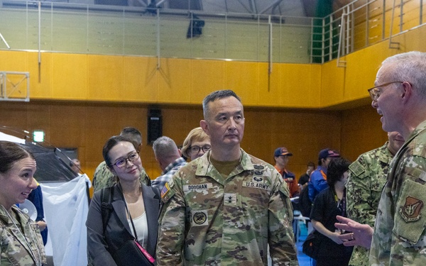 A group of U.S. military personnel in OCP (Operational Camouflage Pattern) uniforms are engaged in conversation inside what appears to be a gymnasium or community center being used as an operational or humanitarian assistance facility. At least three branches appear to be represented, including U.S. Army and U.S. Air Force personnel. A civilian woman in business attire is also present among the group, suggesting a joint civil-military coordination meeting.