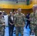A group of U.S. military personnel in OCP (Operational Camouflage Pattern) uniforms are engaged in conversation inside what appears to be a gymnasium or community center being used as an operational or humanitarian assistance facility. At least three branches appear to be represented, including U.S. Army and U.S. Air Force personnel. A civilian woman in business attire is also present among the group, suggesting a joint civil-military coordination meeting.
