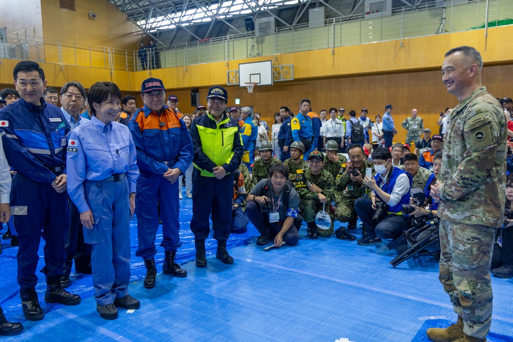 A formal gathering inside a gymnasium or indoor sports facility, showing a U.S. military officer in MultiCam uniform addressing a group of Japanese officials and emergency response personnel. The officials are wearing blue emergency management uniforms with Japanese flag patches, high-visibility vests, and emergency response caps. Photographers and media personnel are crouched in the foreground documenting the event.