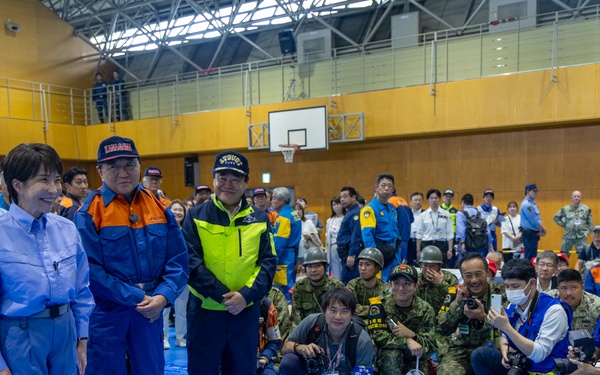 A formal gathering inside a gymnasium or indoor sports facility, showing a U.S. military officer in MultiCam uniform addressing a group of Japanese officials and emergency response personnel. The officials are wearing blue emergency management uniforms with Japanese flag patches, high-visibility vests, and emergency response caps. Photographers and media personnel are crouched in the foreground documenting the event.