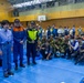 A formal gathering inside a gymnasium or indoor sports facility, showing a U.S. military officer in MultiCam uniform addressing a group of Japanese officials and emergency response personnel. The officials are wearing blue emergency management uniforms with Japanese flag patches, high-visibility vests, and emergency response caps. Photographers and media personnel are crouched in the foreground documenting the event.