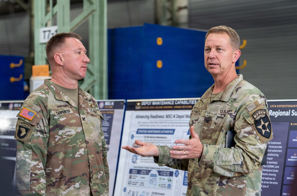 Two U.S. Army officers in OCP (Operational Camouflage Pattern) uniforms are engaged in conversation inside what appears to be a military maintenance depot or hangar. The officer on the right, identified by nametape as 'SCHUCK,' is gesturing and speaking, while the officer on the left, identified as 'CARPENTER,' listens. A briefing board titled 'Depot Maintenance Capabilities' and 'Advancing Readiness: MSC-K Depot Ma[intenance]' is visible in the background.