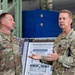 Two U.S. Army officers in OCP (Operational Camouflage Pattern) uniforms are engaged in conversation inside what appears to be a military maintenance depot or hangar. The officer on the right, identified by nametape as 'SCHUCK,' is gesturing and speaking, while the officer on the left, identified as 'CARPENTER,' listens. A briefing board titled 'Depot Maintenance Capabilities' and 'Advancing Readiness: MSC-K Depot Ma[intenance]' is visible in the background.