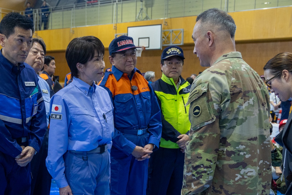 A meeting is taking place inside what appears to be a gymnasium or sports facility between a U.S. military soldier in OCP (Operational Camouflage Pattern) uniform and several Japanese officials wearing blue and orange emergency/government uniforms. The Japanese officials display a Japanese flag patch and kanji text on their uniforms, suggesting government or disaster response roles. The setting and attire indicate a bilateral coordination meeting, likely related to disaster relief or emergency response operations.