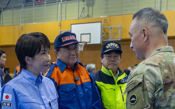 A meeting is taking place inside what appears to be a gymnasium or sports facility between a U.S. military soldier in OCP (Operational Camouflage Pattern) uniform and several Japanese officials wearing blue and orange emergency/government uniforms. The Japanese officials display a Japanese flag patch and kanji text on their uniforms, suggesting government or disaster response roles. The setting and attire indicate a bilateral coordination meeting, likely related to disaster relief or emergency response operations.