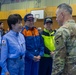 A meeting is taking place inside what appears to be a gymnasium or sports facility between a U.S. military soldier in OCP (Operational Camouflage Pattern) uniform and several Japanese officials wearing blue and orange emergency/government uniforms. The Japanese officials display a Japanese flag patch and kanji text on their uniforms, suggesting government or disaster response roles. The setting and attire indicate a bilateral coordination meeting, likely related to disaster relief or emergency response operations.