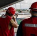 Two workers wearing red hard hats and red safety vests are positioned on an elevated platform, likely aboard a ship or industrial structure at a port facility. One worker is aiming a line-throwing gun (rocket line apparatus), which is standard maritime safety equipment used to send lines between vessels or to shore. The background shows a port environment with parked vehicles, cranes, and industrial infrastructure.
