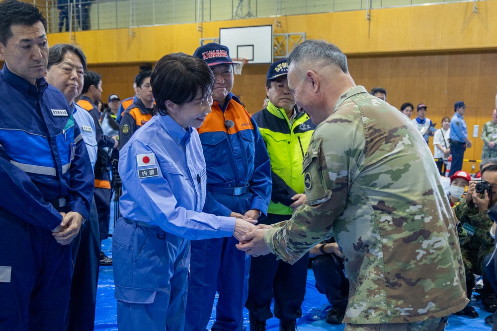 A formal meeting or greeting is taking place inside what appears to be a gymnasium or community center being used as an emergency operations or disaster response center. A person wearing a light blue Japanese government uniform with a Japanese flag patch and '内閣' (Cabinet Office) insignia is shaking hands with a U.S. military personnel in OCP (Operational Camouflage Pattern) uniform. The setting and attire suggest this is a disaster relief coordination meeting, likely in Japan.