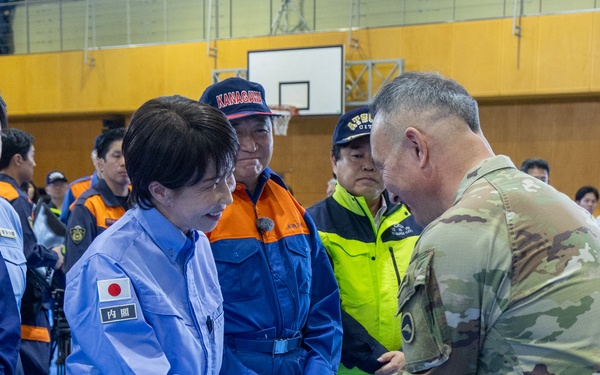 A formal meeting or greeting is taking place inside what appears to be a gymnasium or community center being used as an emergency operations or disaster response center. A person wearing a light blue Japanese government uniform with a Japanese flag patch and '内閣' (Cabinet Office) insignia is shaking hands with a U.S. military personnel in OCP (Operational Camouflage Pattern) uniform. The setting and attire suggest this is a disaster relief coordination meeting, likely in Japan.