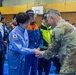 A formal meeting or greeting is taking place inside what appears to be a gymnasium or community center being used as an emergency operations or disaster response center. A person wearing a light blue Japanese government uniform with a Japanese flag patch and '内閣' (Cabinet Office) insignia is shaking hands with a U.S. military personnel in OCP (Operational Camouflage Pattern) uniform. The setting and attire suggest this is a disaster relief coordination meeting, likely in Japan.