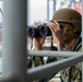 A military service member wearing US Navy digital camouflage (NWU Type III) and a tan/olive MICH combat helmet is using black binoculars to observe something in the distance. The individual has visible tattoos on their hands and neck, and appears to be positioned on or near a ship's railing or scaffolding structure. The background suggests a port or harbor environment with shipping containers visible.