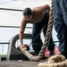 A person in military or naval uniform is working on deck, handling a large mooring line secured around a cleat or bollard. At least two uniformed personnel are visible on what appears to be the deck of a vessel at a port or dock facility. The scene depicts active line-handling operations during a mooring or unmooring evolution.