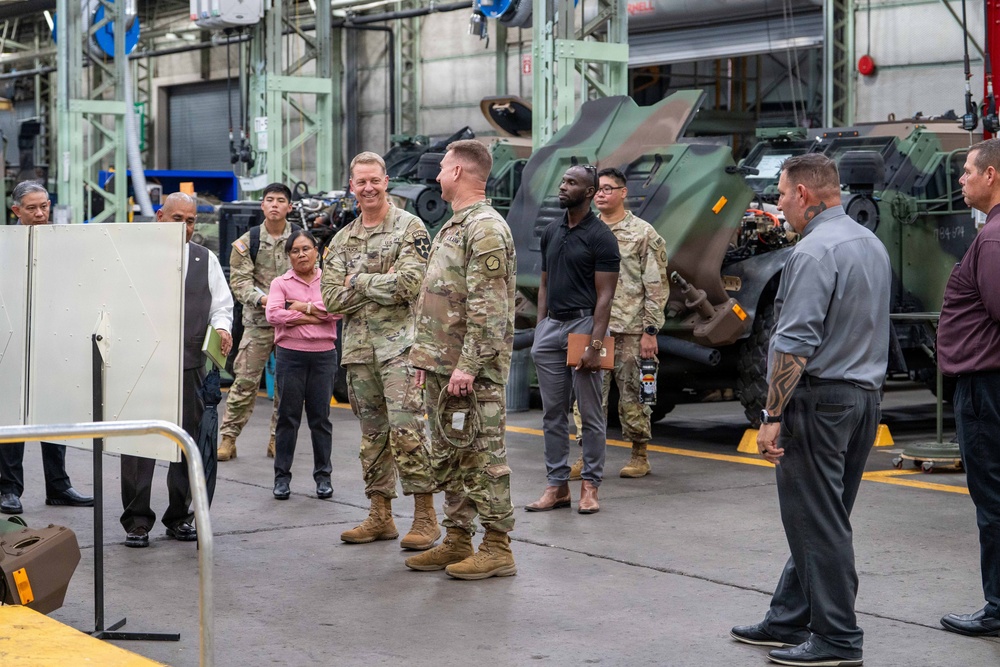 A group of U.S. Army soldiers in OCP (Operational Camouflage Pattern) uniforms and civilian personnel are gathered inside a large military maintenance or depot facility. The setting appears to be an official visit or inspection tour, with military vehicles visible in the background. The atmosphere is informal and conversational among the attendees.