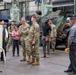A group of U.S. Army soldiers in OCP (Operational Camouflage Pattern) uniforms and civilian personnel are gathered inside a large military maintenance or depot facility. The setting appears to be an official visit or inspection tour, with military vehicles visible in the background. The atmosphere is informal and conversational among the attendees.