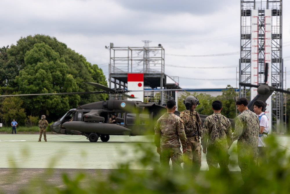 Military personnel from what appear to be multiple services are gathered on a helipad near a dark olive-colored utility helicopter, likely a UH-60 Black Hawk. A Japanese flag emblem is visible on a structure in the background, suggesting this is a joint or bilateral military exercise or visit at a Japanese facility. Personnel in U.S. Army combat uniforms, U.S. Navy working uniforms, and at least one civilian are present.