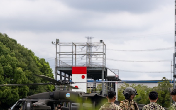 Military personnel from what appear to be multiple services are gathered on a helipad near a dark olive-colored utility helicopter, likely a UH-60 Black Hawk. A Japanese flag emblem is visible on a structure in the background, suggesting this is a joint or bilateral military exercise or visit at a Japanese facility. Personnel in U.S. Army combat uniforms, U.S. Navy working uniforms, and at least one civilian are present.