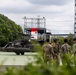 Military personnel from what appear to be multiple services are gathered on a helipad near a dark olive-colored utility helicopter, likely a UH-60 Black Hawk. A Japanese flag emblem is visible on a structure in the background, suggesting this is a joint or bilateral military exercise or visit at a Japanese facility. Personnel in U.S. Army combat uniforms, U.S. Navy working uniforms, and at least one civilian are present.