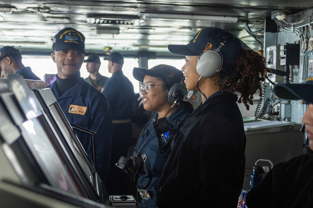 The image shows U.S. Navy personnel on the bridge of what appears to be USS Abraham Lincoln, a naval vessel. Three sailors are prominently featured in the foreground, with one wearing communication headsets and another wearing glasses, all dressed in Navy working uniforms with ship-specific caps. Additional crew members are visible in the background, suggesting an active watch or operational period.